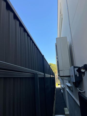 Tall dark corrugated fencing runs beside a white wall with an outdoor air-conditioning unit in a tight passage.