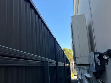 Tall dark corrugated fencing runs beside a white wall with an outdoor air-conditioning unit in a tight passage.