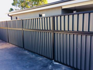Monument panels with a shorter upper infill run along a dark concrete driveway beside a grey house.