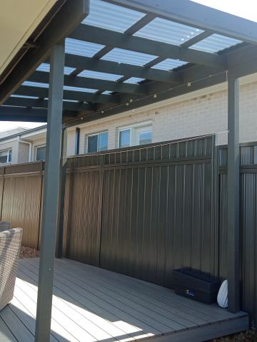 The scene shows a mid grey corrugated steel fence where an upper infill raises the height, concrete paving runs alongside.