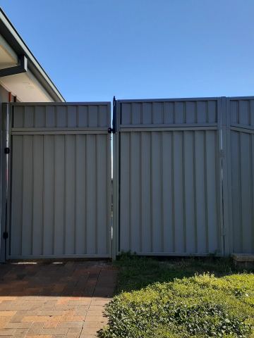 Dark grey pedestrian gate and tapered sheet panels sit on brick paving beside a modern grey and white house.