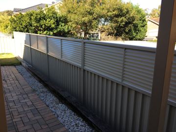 The scene shows a mid grey corrugated steel fence where an upper infill raises the height, residential buildings appear beyond the line.