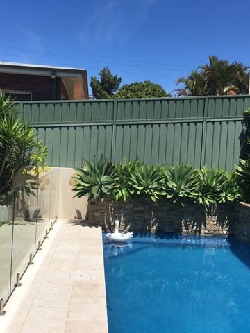 The scene shows a mid grey corrugated steel fence where an upper infill raises the height, residential buildings appear beyond the line.