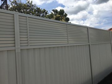 The scene shows a mid grey corrugated steel fence where an upper infill raises the height, concrete paving runs alongside.