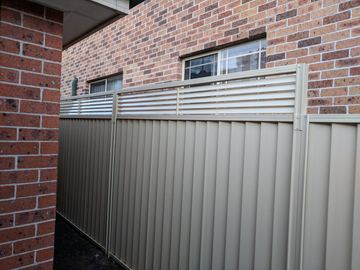 The scene shows a mid grey corrugated steel fence where concrete paving runs alongside.
