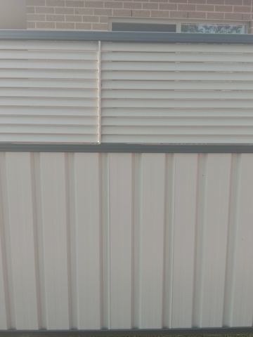The scene shows a light grey corrugated steel fence where concrete paving runs alongside.