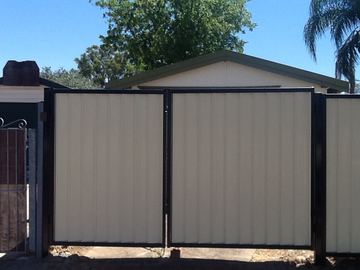 The scene shows a mid grey corrugated steel fence where concrete paving runs alongside.