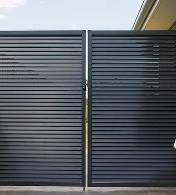 The scene shows a mid grey corrugated steel fence where a gate sits in the run, louvre sections add ventilation, concrete paving runs alongside.