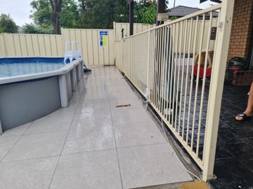 Cream flat-top tubular panels and gate ring a wet tile walk beside cream corrugated boundary fencing with a safety sign.