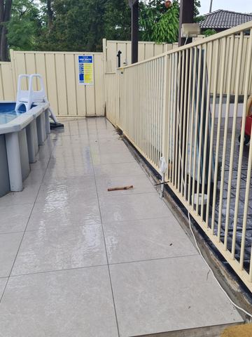 Cream flat-top tubular panels and gate ring a wet tile walk beside cream corrugated boundary fencing with a safety sign.
