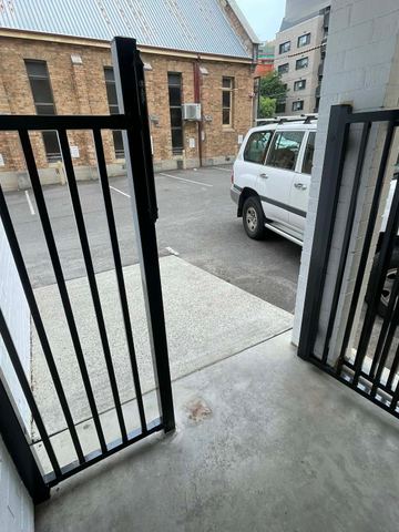 Black flat-top picket gate and panel open from a white brick porch toward an aggregate path and parked cars.