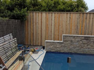 Vertical timber slats on stone tiers stand beside frameless glass pool fencing, tools and brushwood screening.