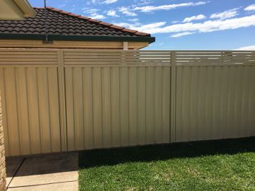 Cream panels with three-line slat tops cast shadows on grass beside a brick house corner under blue sky.