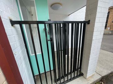 Black picket gate closes a white-brick alcove in front of a mint-green glazed door under a ceiling light.
