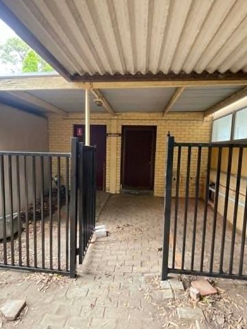 Black picket fence and gate span a brick-paved passage beneath a corrugated awning beside a cream brick building.